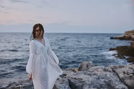 Женщина в белом платье стоит на камнях на берегу океана