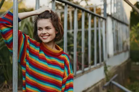 Женщина, портрет молодой женщины, улыбка, красочный свитер, теплица на открытом воздухе, повседневный искренний снимок улыбающейся девушки в полосатом трикотаже, стоящей у стеклянной конструкции…