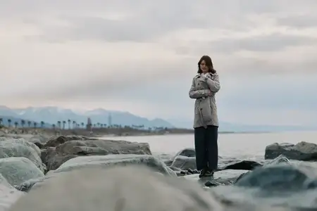 Задумчивая женщина в длинном пальто стоит на больших камнях у моря под облачным небом