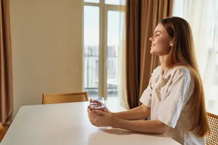 Женщина со стаканом сидит за столом у окна со шторами