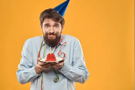 Весёлый мужчина с тортом на жёлтом фоне, на голове у него праздничный колпак в честь дня рождения