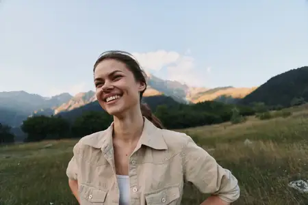 Счастливая женщина стоит на травянистом поле на фоне гор, в повседневной рубашке; естественный солнечный свет освещает её лицо, мирная сцена отдыха на природе