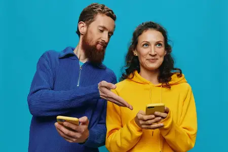 Весёлая пара — женщина и мужчина — с телефонами в руках, криво и радостно улыбаются на синем фоне