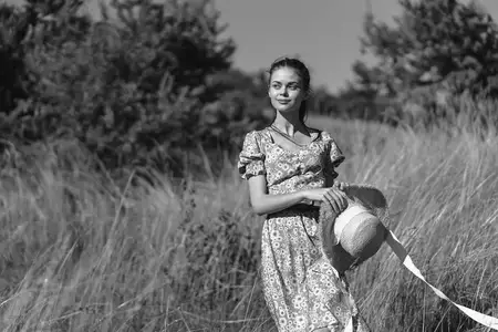 Женщина в элегантном платье и шляпе идет по высокой траве во время модной фотосессии недалеко от сельской местности