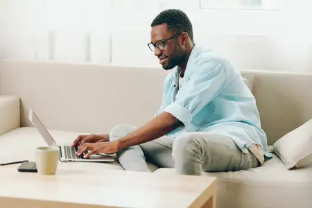 Улыбающийся афроамериканский мужчина работает за ноутбуком в уютном домашнем офисе, разговаривает по телефону и наслаждается онлайн-шопингом