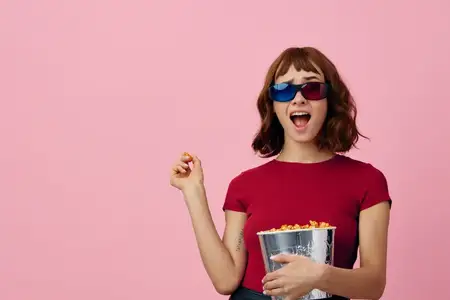 Необычайно счастливая, милая рыжеволосая девушка в красной футболке и 3D-очках с попкорном смотрит фильм, позируя изолированно на розовом фоне в студии
