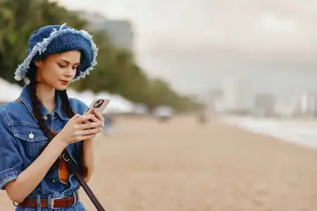 Молодая женщина держит смартфон и наслаждается летним отдыхом на пляже, печатая сообщение с милой улыбкой