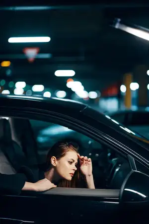 Вертикальная фотография, снятая сбоку ночью: женщина сидит в чёрном автомобиле и смотрит в окно, глядя в боковое зеркало, при этом держит руку возле лица