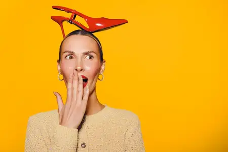Женщина с красной туфлей на голове выглядит удивлённой, прикрывая рот рукой