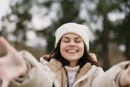 Портрет радостной женщины, тянущейся к камере в заснеженном парке, в теплой куртке и шапке, яркая улыбка и естественный свет, передающие беззаботное настроение