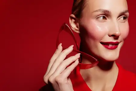 Улыбающаяся женщина с глянцевыми красными губами держит красное украшение в форме сердца возле щеки на красном фоне