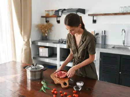Задумчивая женщина на кухне готовит обед из мяса, овощей и специй на деревянном столе на фоне стильной кухни