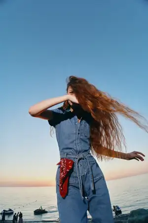 Женщина в стиле лайфстайл, одетая в современный бохо-вестерн деним в уличном стиле, позирует у океана на закате, ее длинные волосы свободно развеваются, а естественные теплые кинематографические…