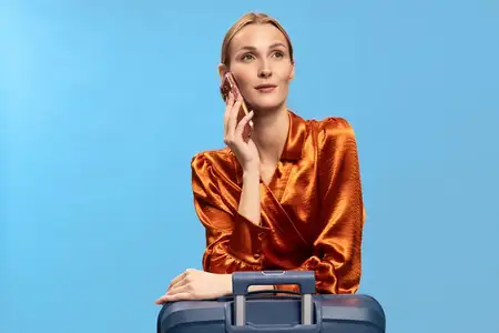 Женщина в блестящей оранжевой рубашке разговаривает по мобильному телефону, опираясь на чемодан