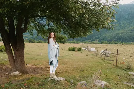 Женщина, укутанная в одеяло, стоит возле дерева на открытом поле на фоне гор