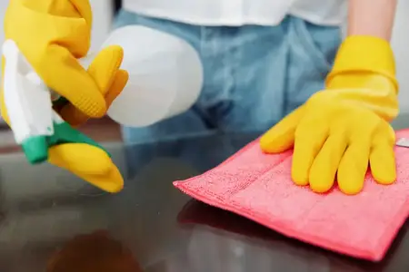 Женщина в жёлтых перчатках протирает стол тряпкой и пульверизатором с чистящим раствором