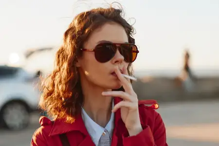 Молодая женщина в солнцезащитных очках и красном пальто курит сигарету на улице в мягком солнечном свете