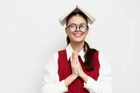Радостная студентка в очках держит книгу на голове, демонстрируя игривое настроение
