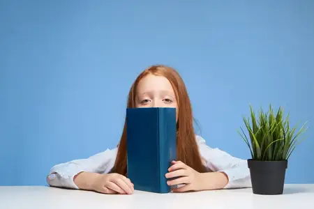 Рыжеволосая девушка сидит за школьной партой с книгой в руках