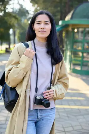 Сцена уличной фотографии: женщина с камерой в парке, повседневная одежда, мягкий свет, задумчивая поза, городская природа, тяга к путешествиям, повседневное повествование через объектив