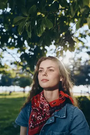Портрет женщины в стиле лайфстайл, одетой в современную богемно-вестерн джинсовую уличную одежду с красной банданой, стоящей на открытом воздухе под густыми зелёными листьями и мягким солнечным…