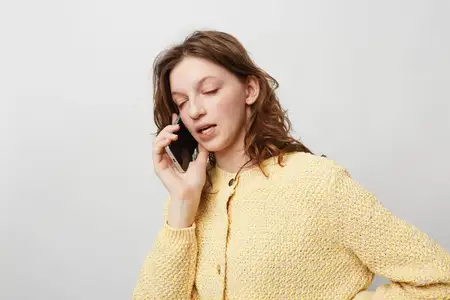 Женщина в желтом свитере прижимает смартфон к уху с закрытыми глазами, демонстрируя задумчивое выражение лица
