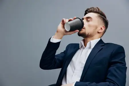 Уставший бизнесмен с чашкой кофе работает за компьютером на сером фоне