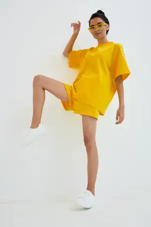 Стильная молодая женщина в ярко-желтой oversized футболке и шортах принимает динамичную позу на однотонном белом фоне, дополненная желтыми солнцезащитными очками и белыми кроссовками