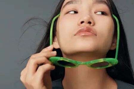 Крупный план женщины с тёмными волосами, поднимающей ярко-зелёные солнцезащитные очки рукой и смотрящей в сторону