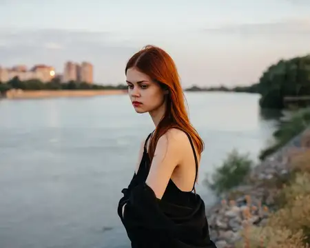 Рыжеволосая женщина в черном платье стоит у реки на природе