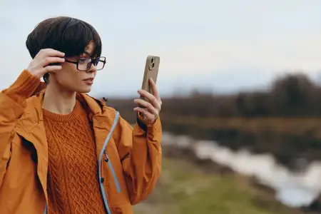 Портрет женщины на открытом воздухе у воды: держит смартфон для селфи, очки и теплая куртка, повседневный лайфстайл, естественный свет, общение, современные технологии