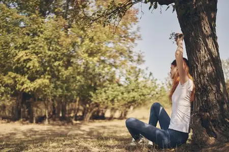 Счастливая женщина в джинсах и футболке сидит у дерева, подняв руки вверх