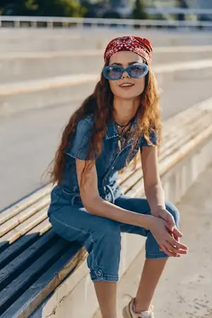 Улыбающаяся молодая женщина в денимовом наряде с красной повязкой на голове и солнцезащитными очками отдыхает на деревянной скамейке на улице под ярким солнечным светом, наслаждаясь теплой погодой и…