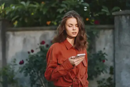 Уличный портрет женщины, использующей смартфон в саду, демонстрирующий современную моду, спокойное настроение и аутентичный образ жизни в яркой обстановке