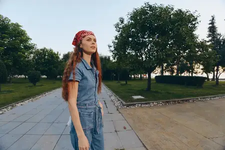 Молодая женщина, ведущая активный образ жизни, одетая в современный бохо-вестерн джинсовый уличный стиль с красной банданой, уверенно идёт по парковой дорожке в окружении пышных деревьев и мягких…