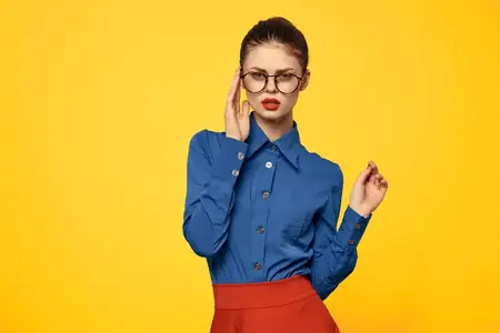 Женщина в голубой рубашке и красной юбке, очки на лице, уверенный взгляд, портрет, желтый фон, обрезанный вид
