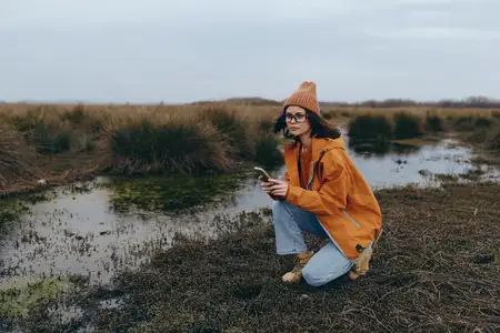 Осенняя фотосцена на природе в стиле лайфстайл: женщина стоит на коленях у реки, одетая в куртку, шарф и очки, повседневные джинсы и вязаную шапку, с умиротворённой улыбкой, тихий день у воды