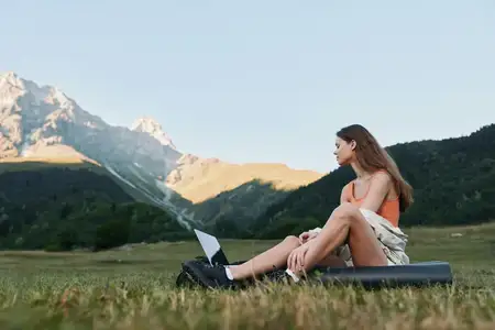 Outdoor-сцена показывает женщину, отдыхающую с ноутбуком на лугу, на фоне возвышающихся гор