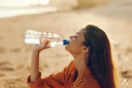 Одинокая женщина на песчаном пляже пьет из бутылки с водой, в то время как солнечный свет отражается от прозрачного пластика