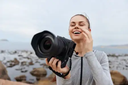 Радостная женщина-фотограф улыбается, снимая камерой живописные прибрежные пейзажи, демонстрируя страсть к фотографии и связь с природой