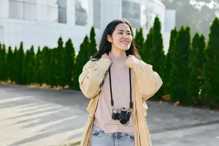 Женщина-фотограф в повседневной одежде на открытом воздухе: бежевая куртка и мягкий свитер, фотоаппарат висит на шее, наслаждается солнечным днем рядом с зелеными деревьями и современным зданием