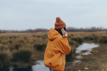 Женщина отдыхает на природе в дождевике и шапке на фоне реки и деревенского пейзажа