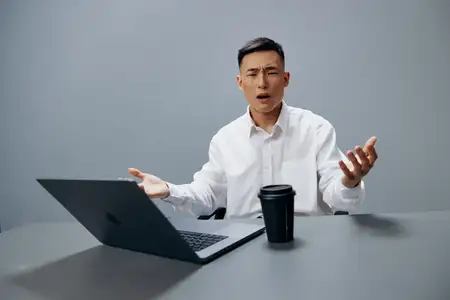 Бизнесмен испытывает усталость и эмоции, работая за ноутбуком с чашкой кофе на рабочем столе на сером фоне
