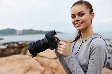 Счастливая молодая женщина-фотограф в серой толстовке с камерой в руках у океана, что отражает любовь к фотографии и приключениям на открытом воздухе