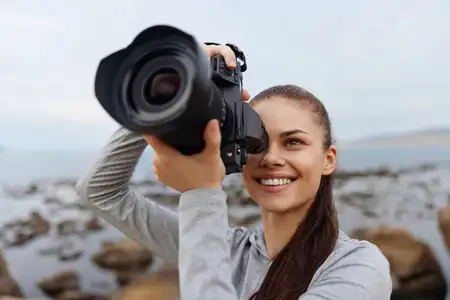 Веселая молодая женщина с длинными волосами наслаждается фотосъемкой на пляже, демонстрируя свою страсть к запечатлению ярких моментов природы с помощью профессиональной камеры