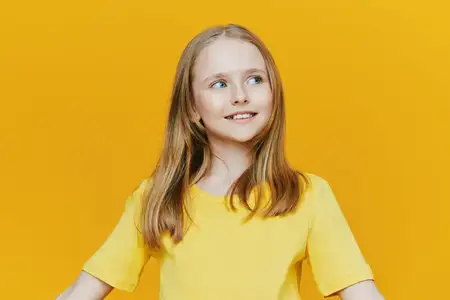 Весёлая маленькая девочка с длинными волосами стоит и улыбается на ярком жёлтом фоне, одетая в подходящую по цвету жёлтую футболку