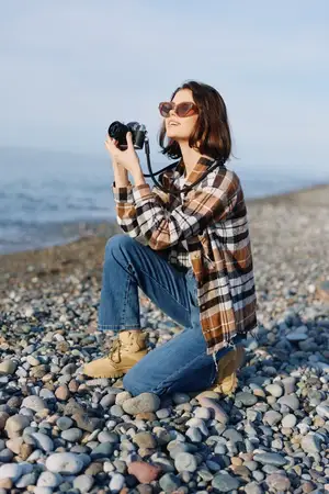 Сцена уличной фотографии: женщина с камерой на галечном берегу, одетая в клетчатую рубашку и джинсы, присела и снимает морской пейзаж с сосредоточенностью и стилем