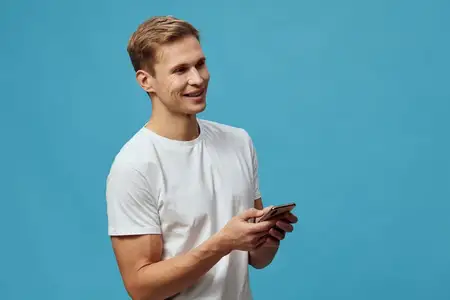 Улыбающийся молодой человек в белой футболке держит смартфон и взаимодействует с приложением чат-бота с искусственным интеллектом в непринужденной позе на синем фоне
