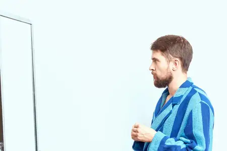 Мужчина в синем халате рассматривает себя в зеркале в светлой комнате, обрезанный вид
