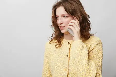 Женщина с вьющимися волосами в жёлтом трикотаже прижимает телефон к уху, глядя в сторону